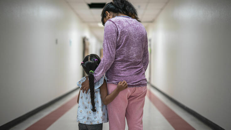 A Mexican mother wearing pink pants and a purple shirt stands with her arms around her young daughter, who has a long braid, in a hallway. They are asylum seekers in an IRC welcome center.
