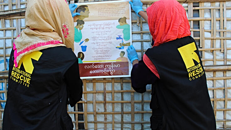 Two women post coronavirus information on the wall of a bamboo shelter in their roles as IRC Community health workers