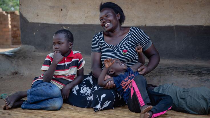 Jackie sits on the ground smiling with her two sons. The older is leaning against her and looking into the distance and the younger is lying on his back on his mothers lap, laughing.