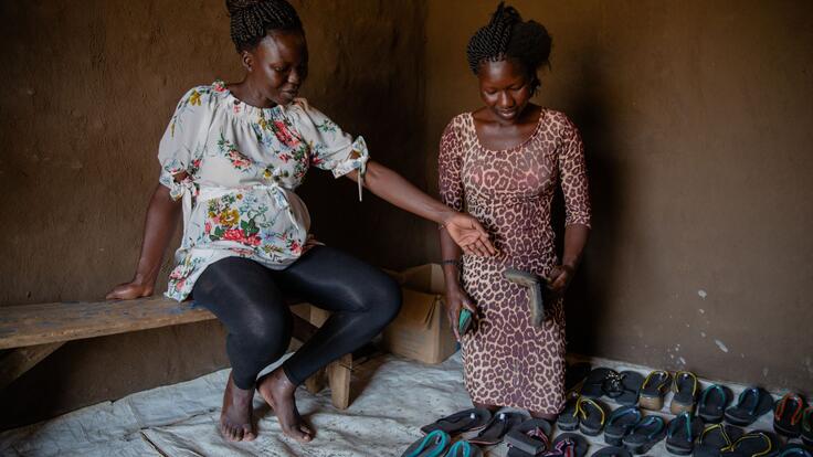 Grace sits on a bench next to a refugee named Vivian, who is kneeling next to shoes arranged neatly on the ground. She is holding one of the shoes and looking down at them.