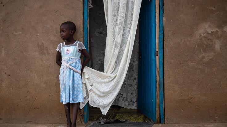 Foni Grace’s six-year-old daughter, Nancy, stands outside their home. She is looking to the side and holding the edge of a white curtain.