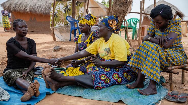 Jemimah and other Togoleta members counsel a woman named Cecilia. Jemimah is sitting on a blanket on the ground next to Cecilia, holding her hand. The other two women are sitting on chairs nearby.