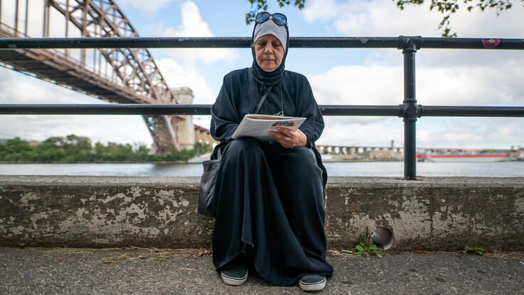Maha sits on the curb next a river while looking at the book she uses to study for her citizenship. There is a bridge behind her in the distance.